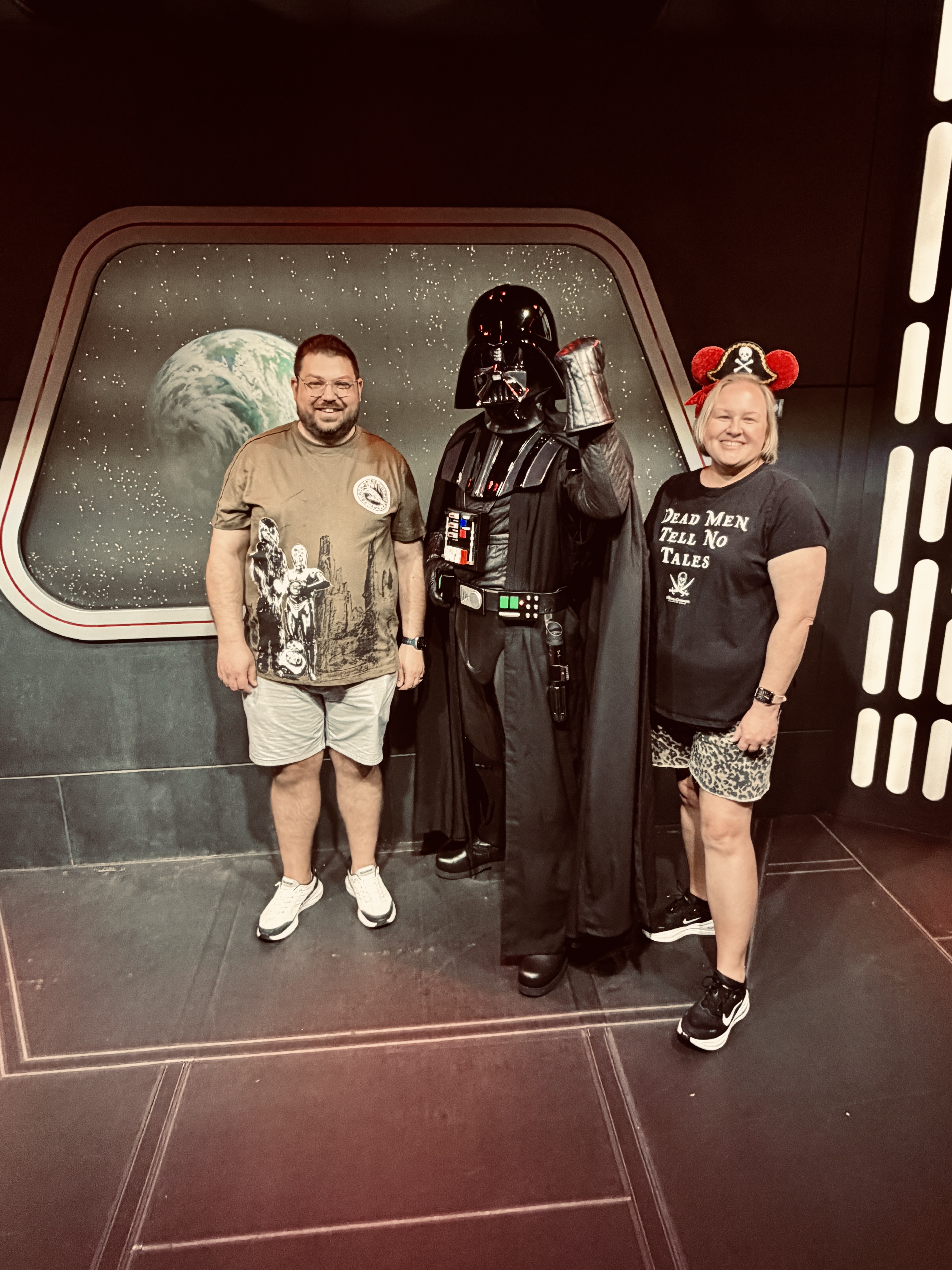 Two guests posing with Darth Vader in front of a starship window.