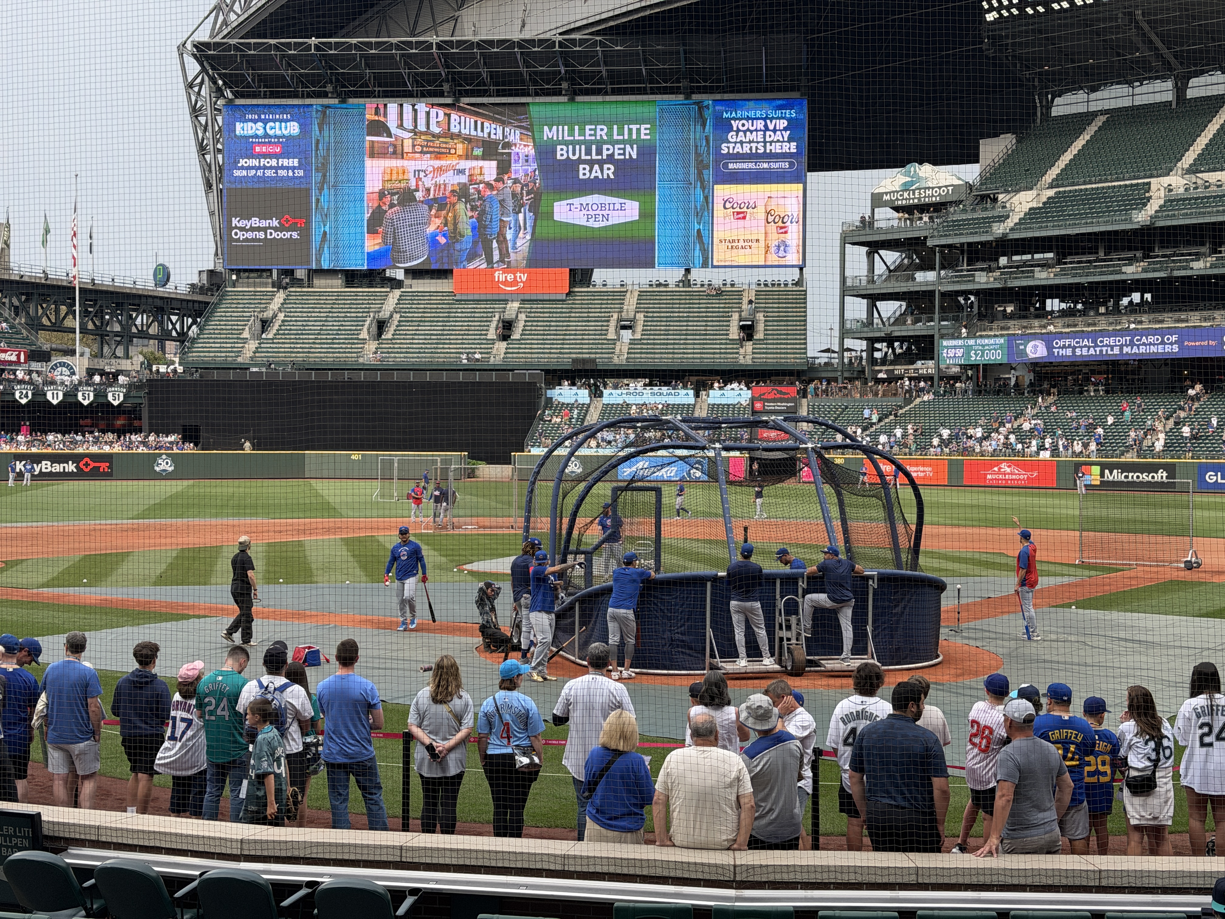Players taking batting practice in the cage before the game, fans lined along the rail.