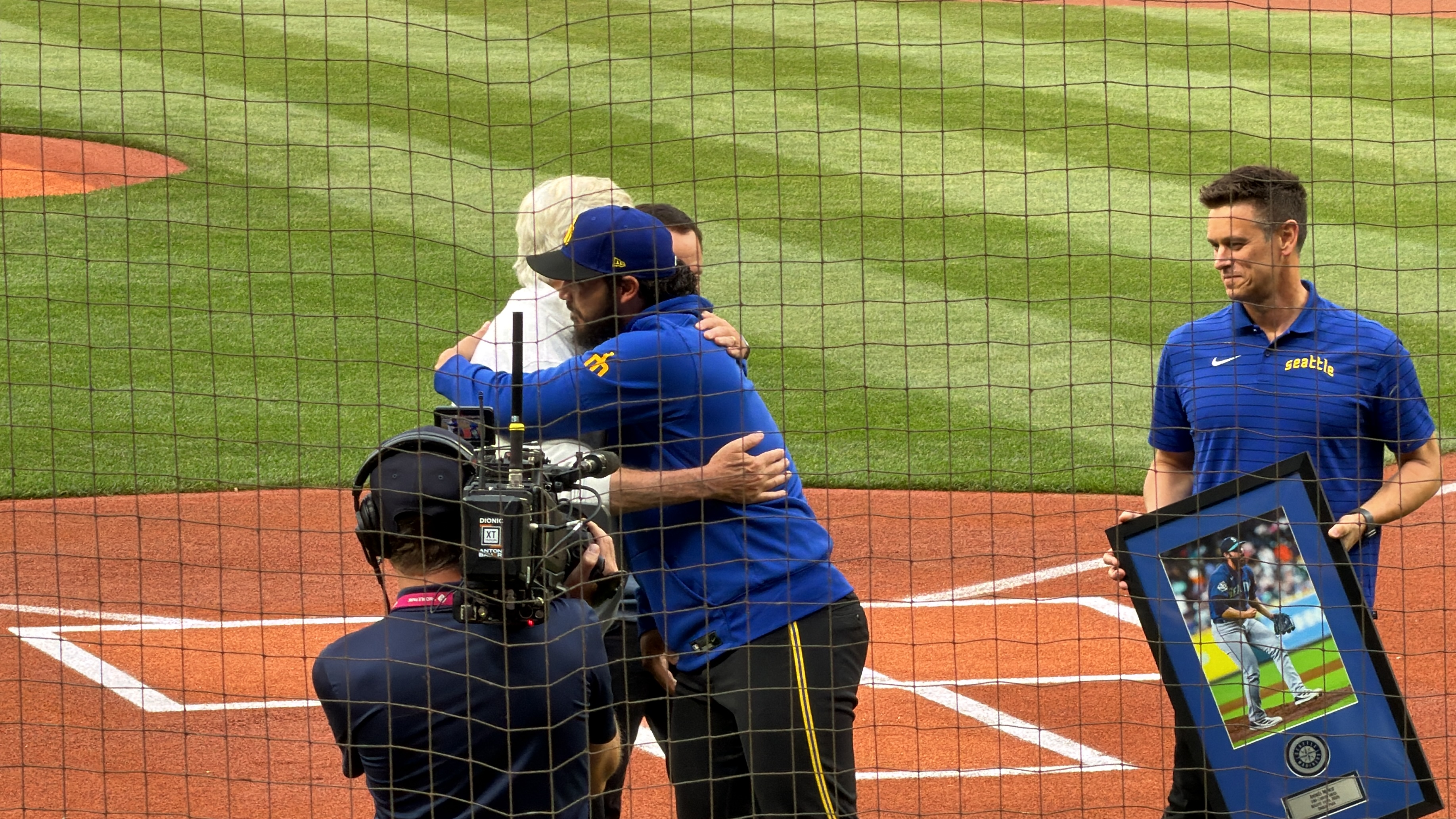 Two people hugging at home plate while another holds a framed photo.