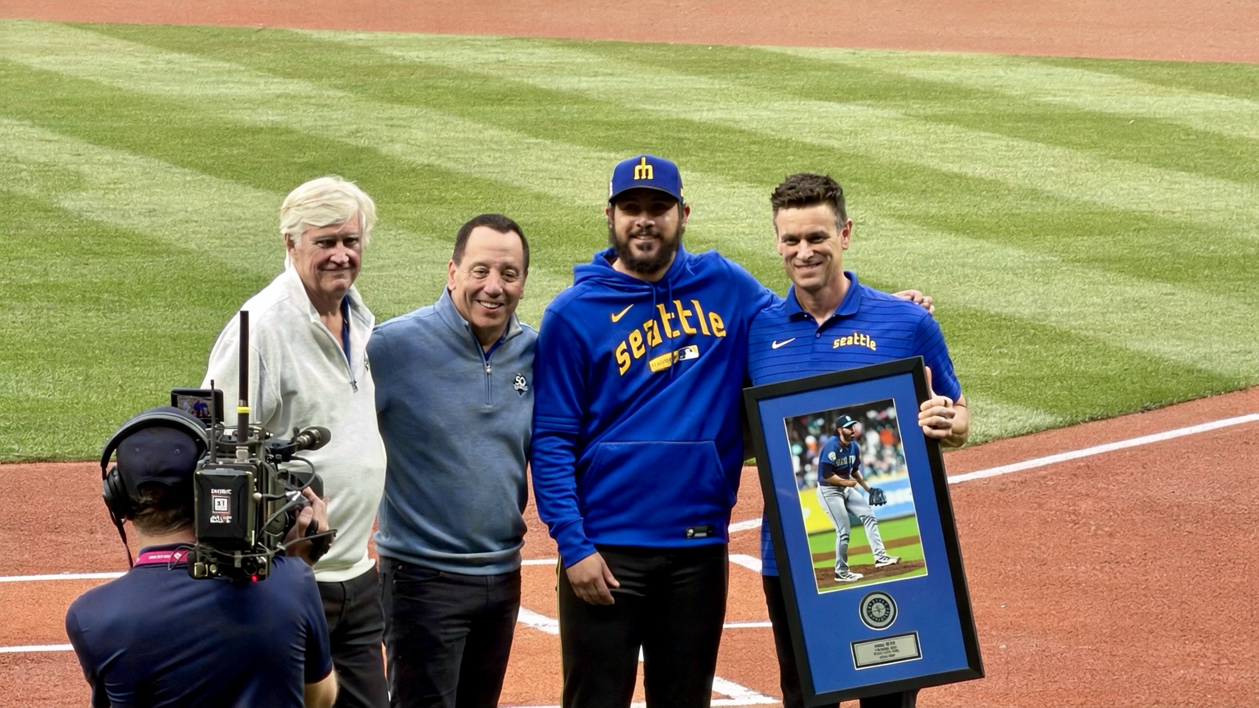 Four people posing at home plate, one holding a framed photo, with a TV camera in front.